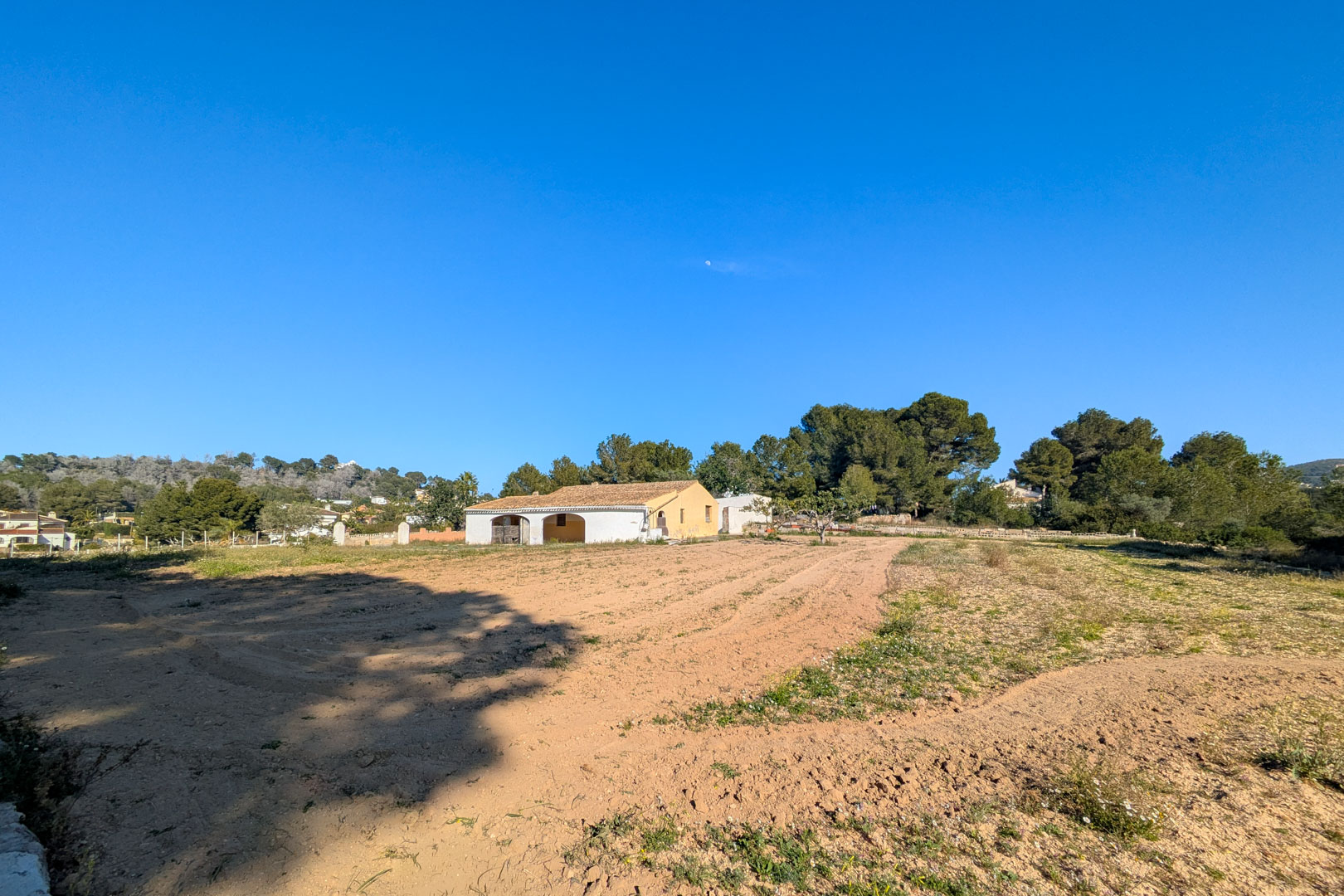 Vista exterior de la finca tradicional con porche de arcos, ubicada en una amplia parcela rústica llana rodeada de pinos y vegetación autóctona en Jávea.