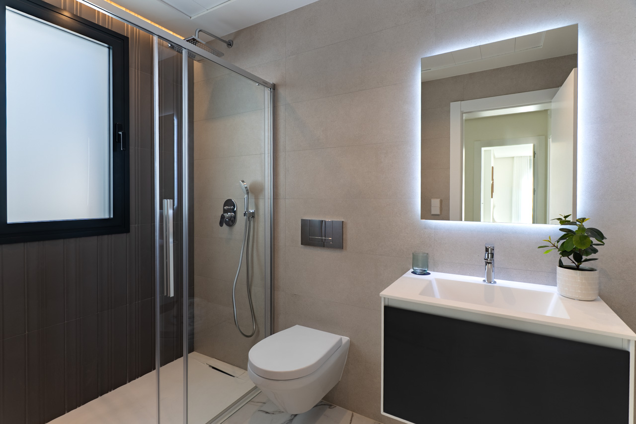 Baño de diseño contemporáneo con ducha de obra, mampara de cristal, inodoro suspendido y mueble de lavabo con espejo retroiluminado sobre gres cerámico en tonos tierra.