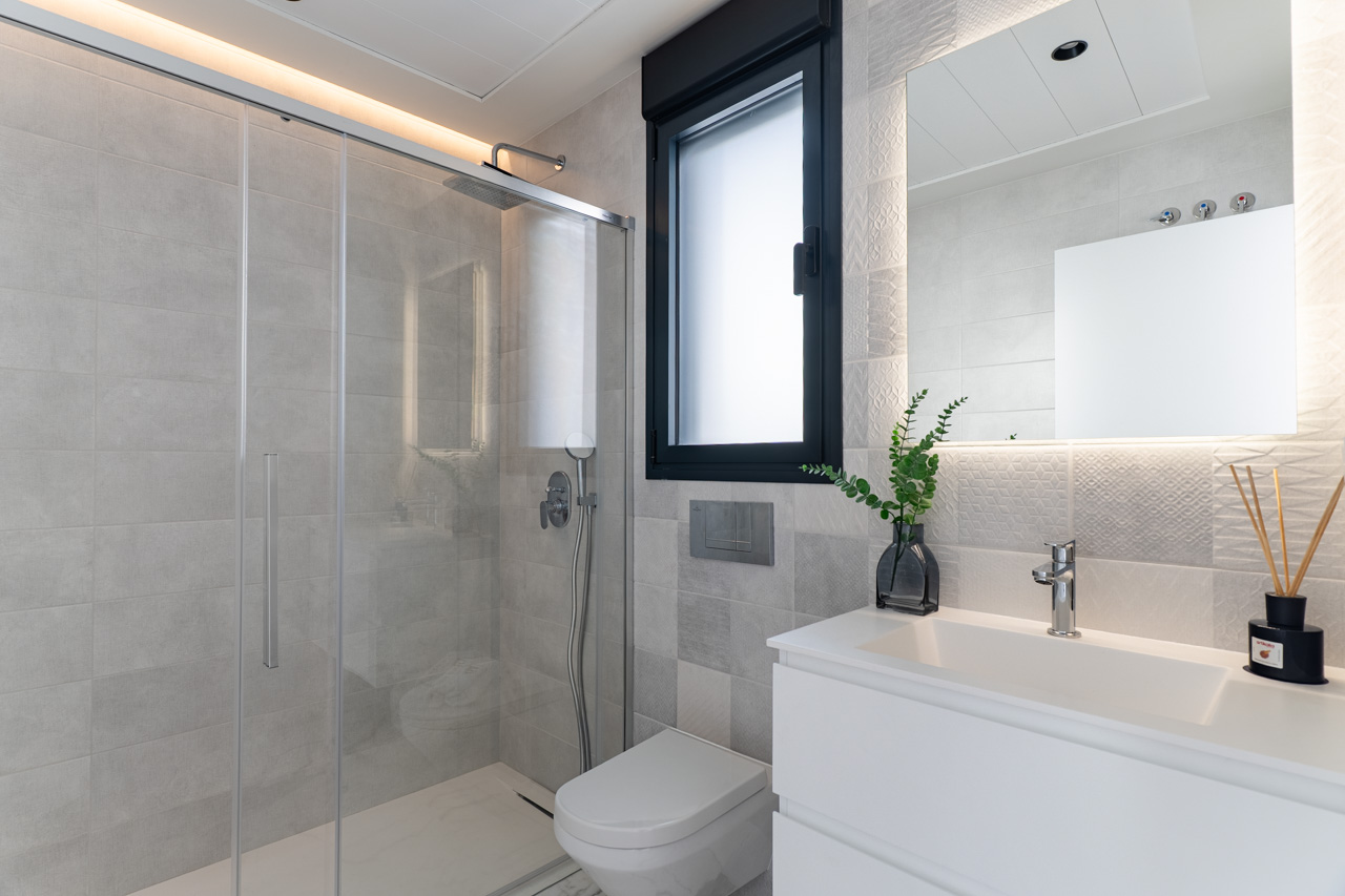 Baño moderno con ducha de obra, mampara de cristal, azulejos texturizados en tonos grises, lavabo blanco y espejo retroiluminado con luz LED indirecta.