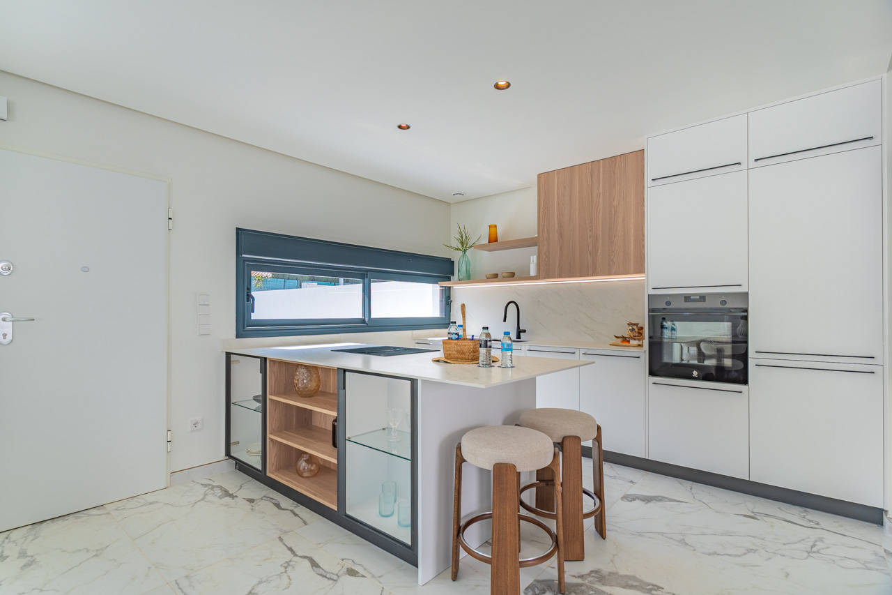 Cocina moderna de concepto abierto con isla central, encimeras blancas y acabados en madera. Incluye electrodomésticos integrados y suelos de porcelánico con efecto mármol.