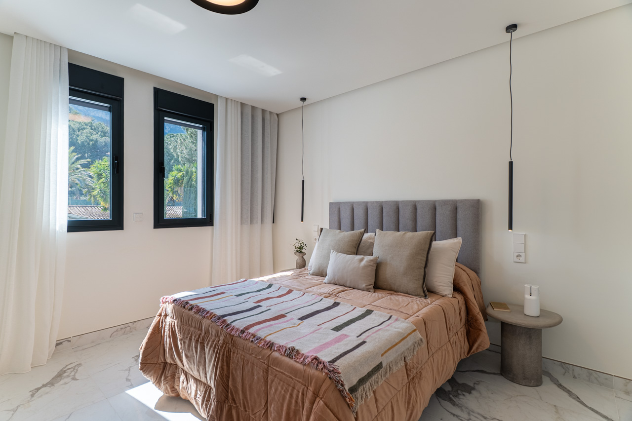 Dormitorio doble con suelo de gres porcelánico efecto mármol, cabecero tapizado en gris y ventanales con marcos negros que ofrecen vistas al entorno natural de Dénia.