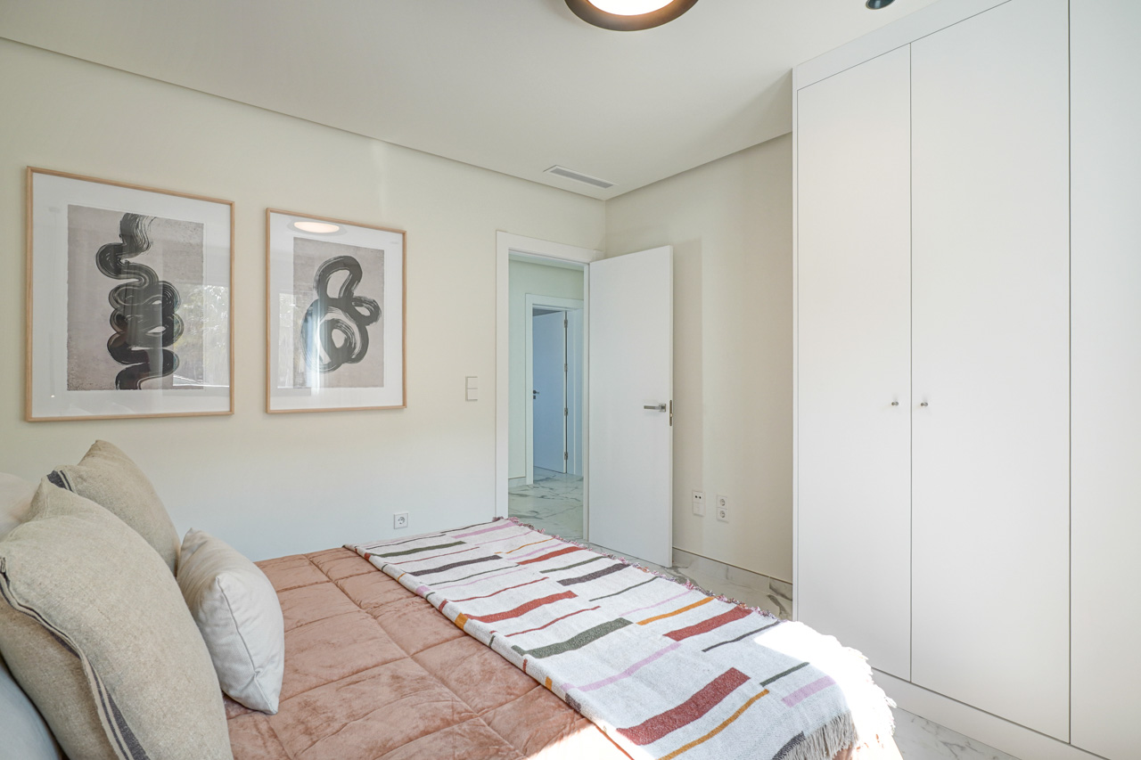 Dormitorio luminoso con armarios empotrados de suelo a techo en blanco, suelos de gres efecto mármol y decoración minimalista con cuadros abstractos.