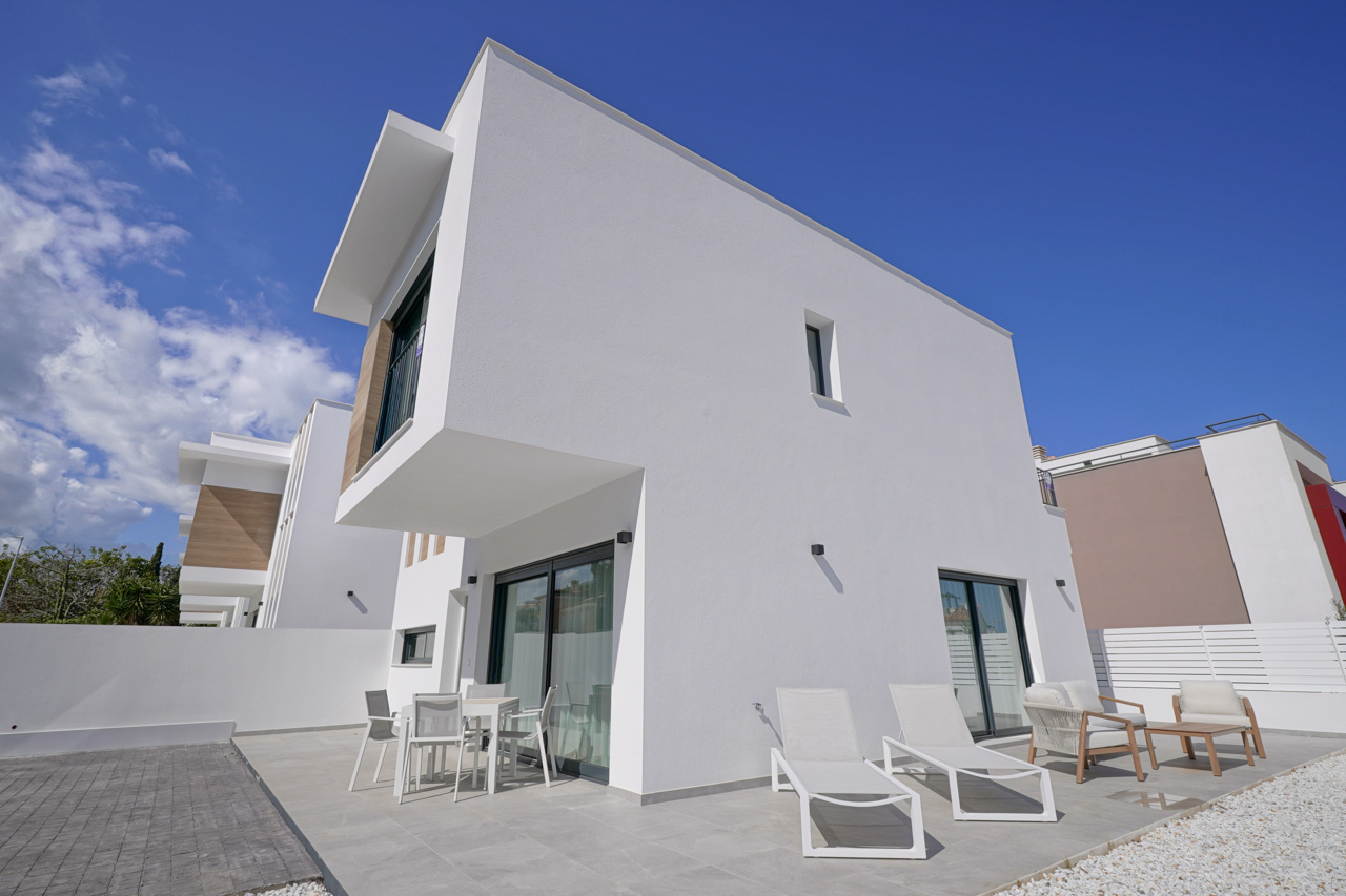 Fachada exterior de diseño contemporáneo con volúmenes blancos, terraza pavimentada con mobiliario de exterior y zona de aparcamiento privada.