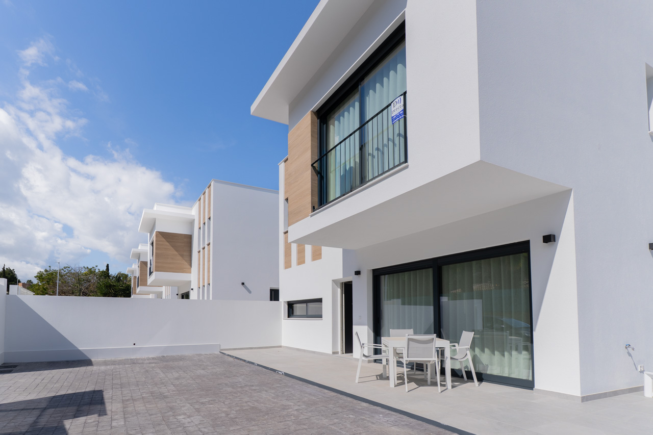 Fachada moderna de líneas rectas con paneles de efecto madera, terraza pavimentada y zona de comedor exterior junto a grandes ventanales correderos.