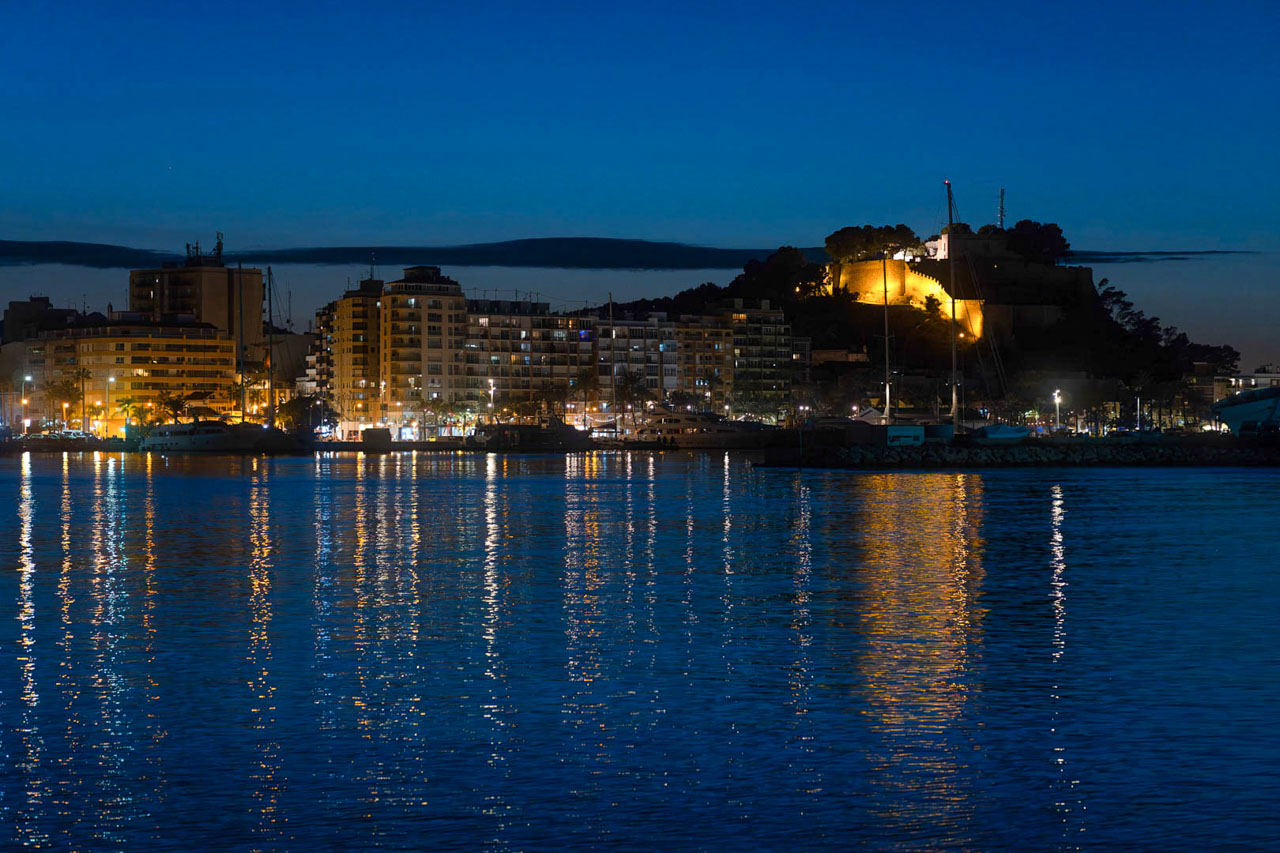 Vista nocturna del puerto de Dénia con el castillo iluminado sobre la colina y los reflejos de las luces de la ciudad sobre el agua del Mediterráneo.