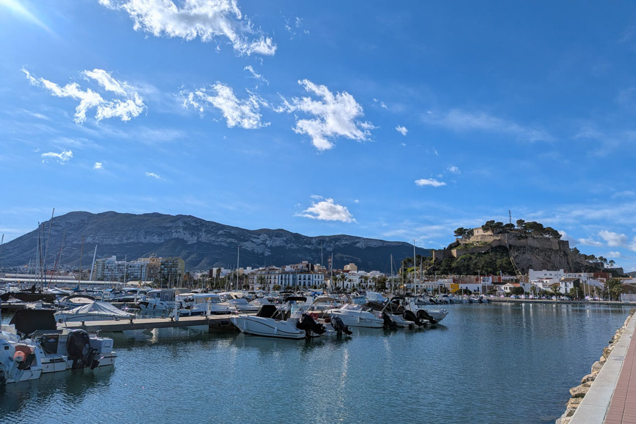Vista panorámica del puerto deportivo de Dénia con el histórico castillo y el macizo del Montgó al fondo bajo un cielo despejado.