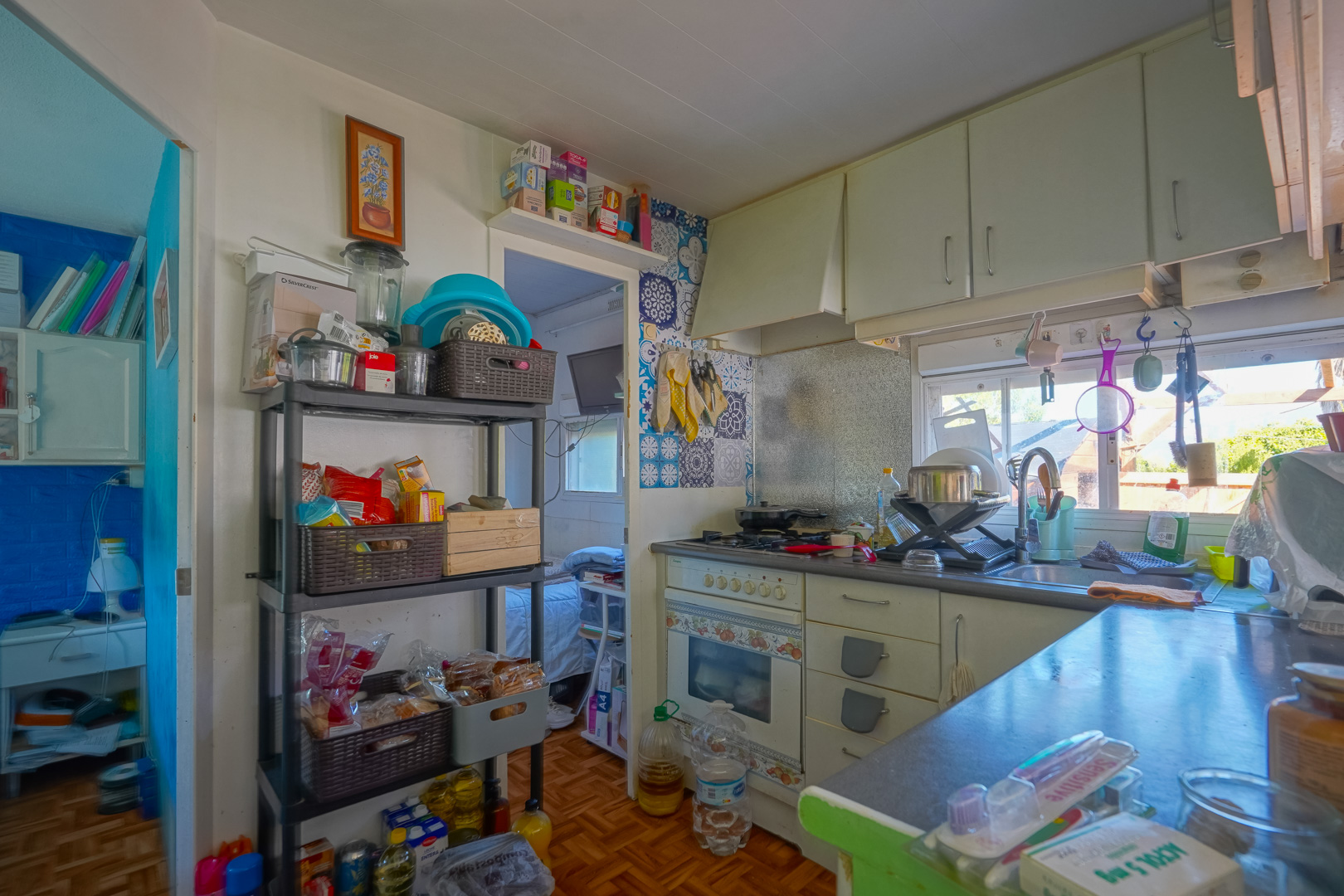 Cocina compacta de la vivienda auxiliar con armarios blancos, zona de cocción con azulejos decorativos y estanterías de almacenaje junto a un ventanal luminoso.