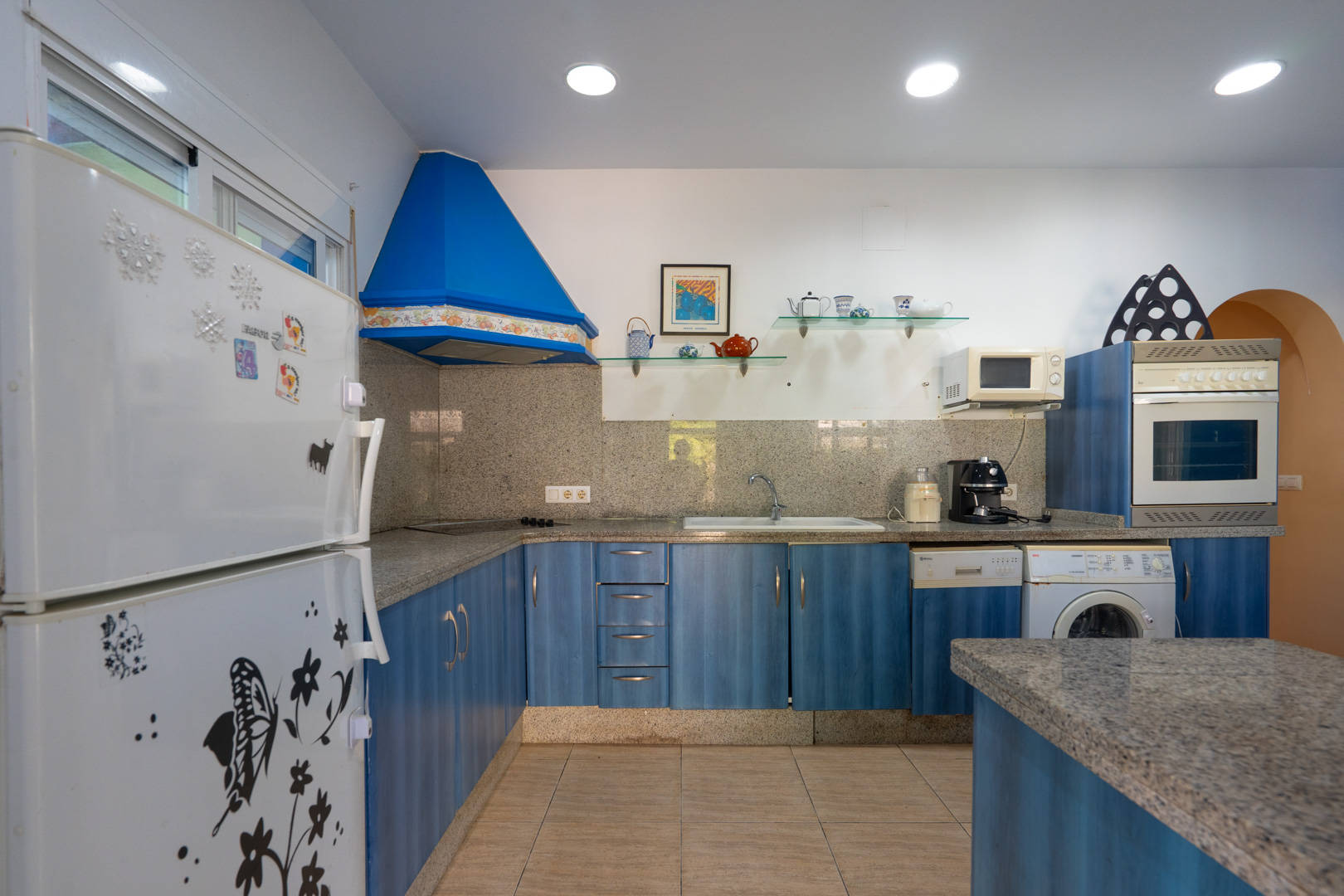 Cocina funcional con armarios azules, encimera de granito gris y electrodomésticos integrados. Incluye campana extractora azul, estantes de cristal y suelos cerámicos.
