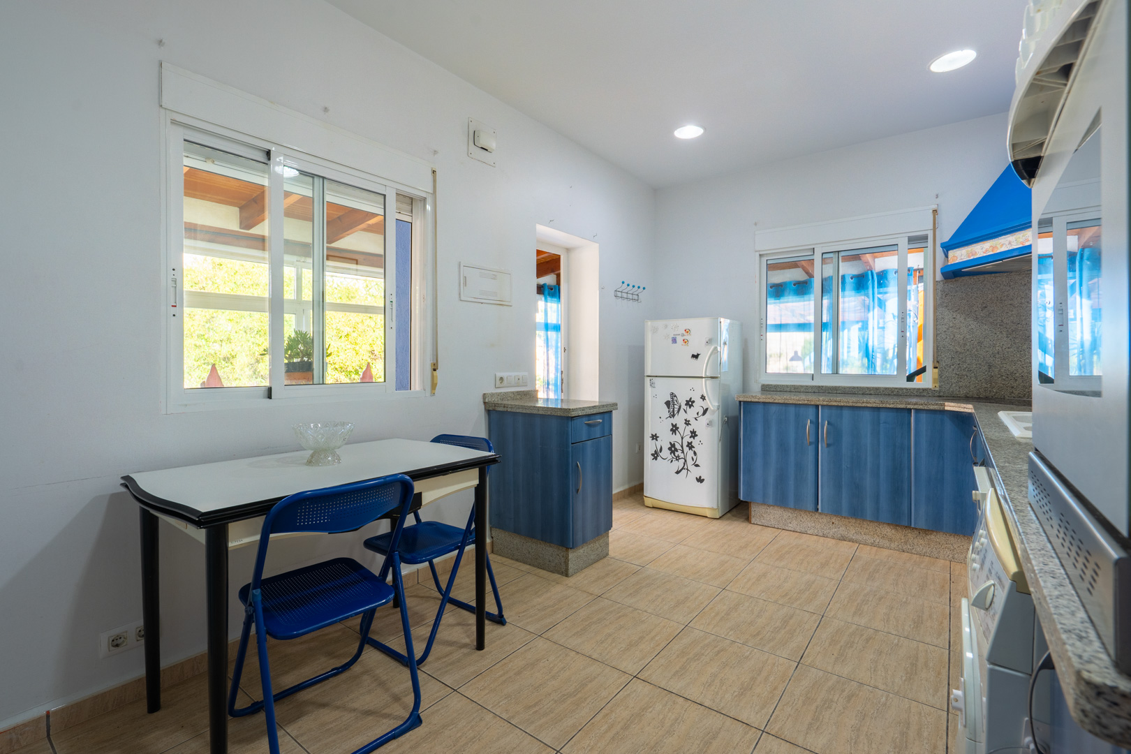 Cocina funcional con armarios azules, encimeras de granito y zona de comedor integrada. Dispone de iluminación empotrada y vistas al exterior a través de ventanas de aluminio.