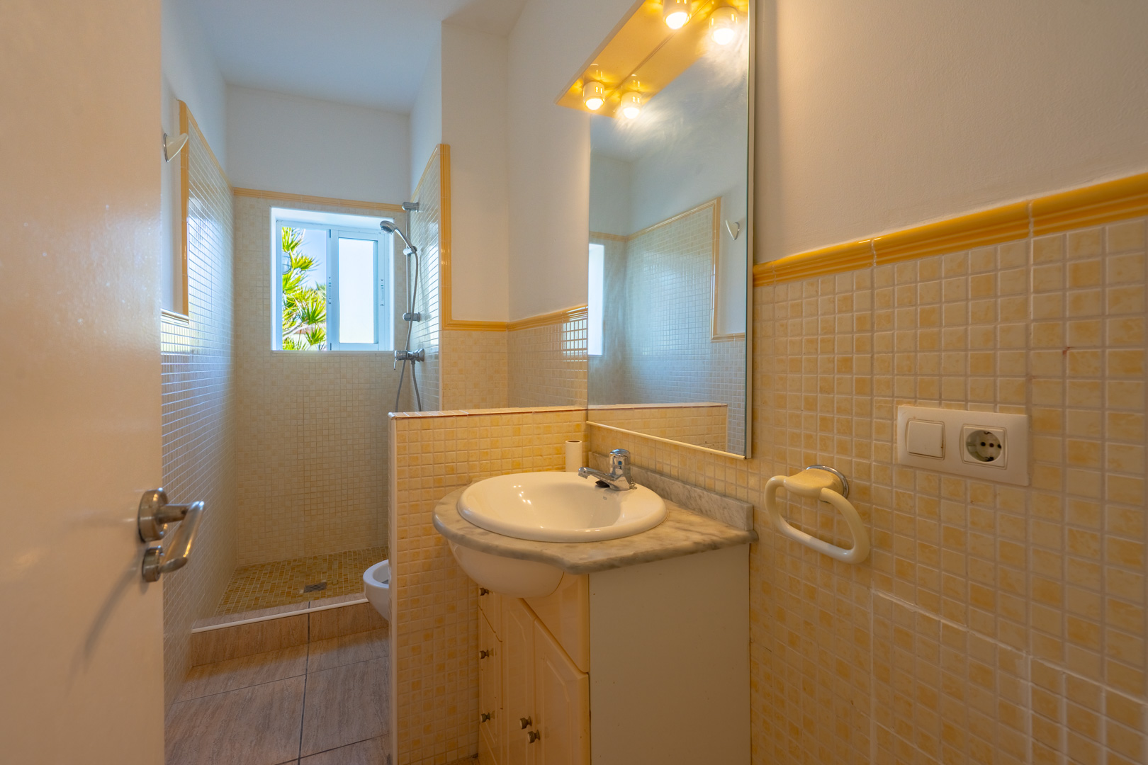 Cuarto de baño con ducha de mosaico, encimera de mármol y espejo con iluminación integrada. Una ventana aporta luz natural y vistas al exterior.
