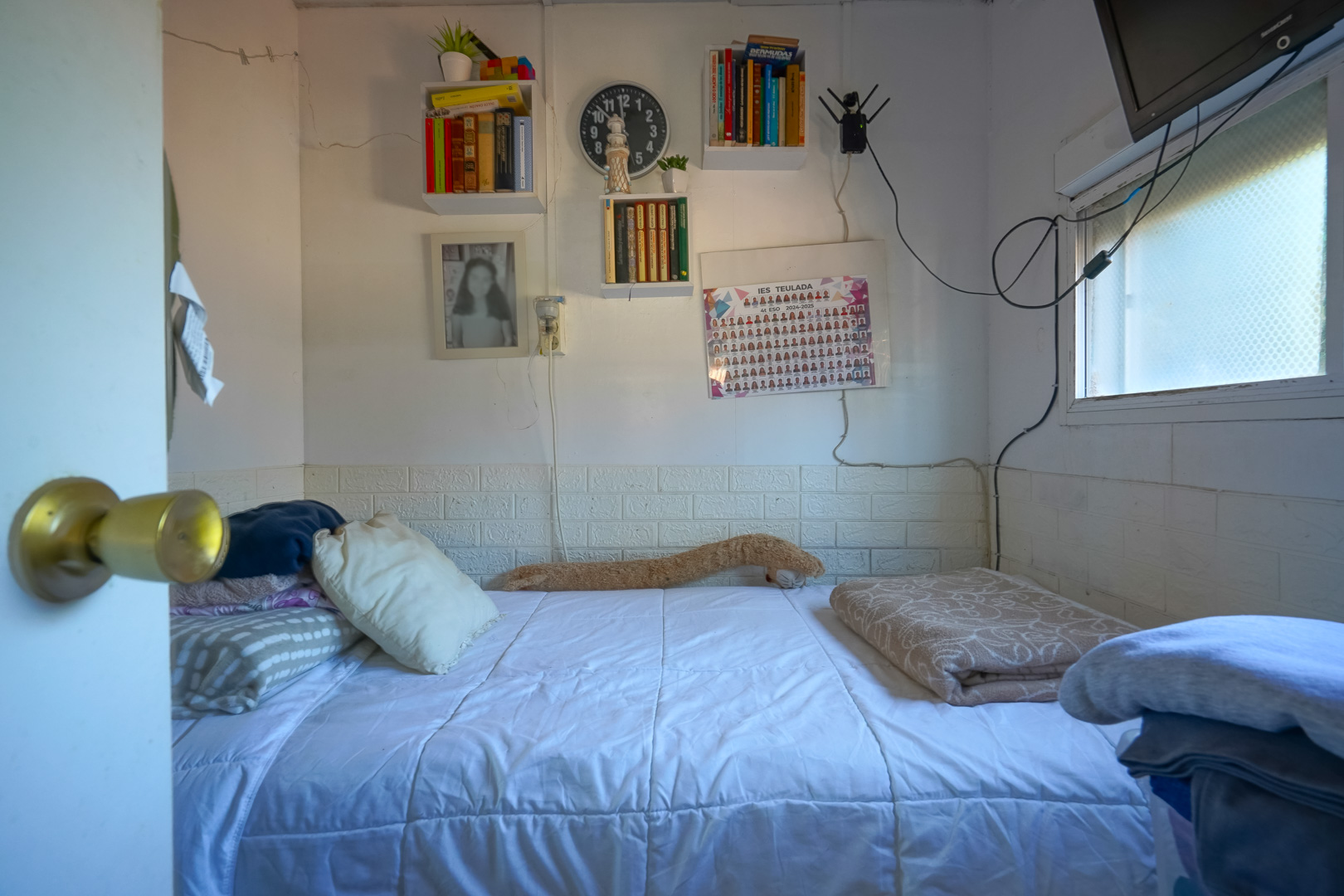 Dormitorio compacto con cama individual, estantes de pared para libros y ventana lateral. Presenta un zócalo decorativo con efecto de ladrillo blanco.