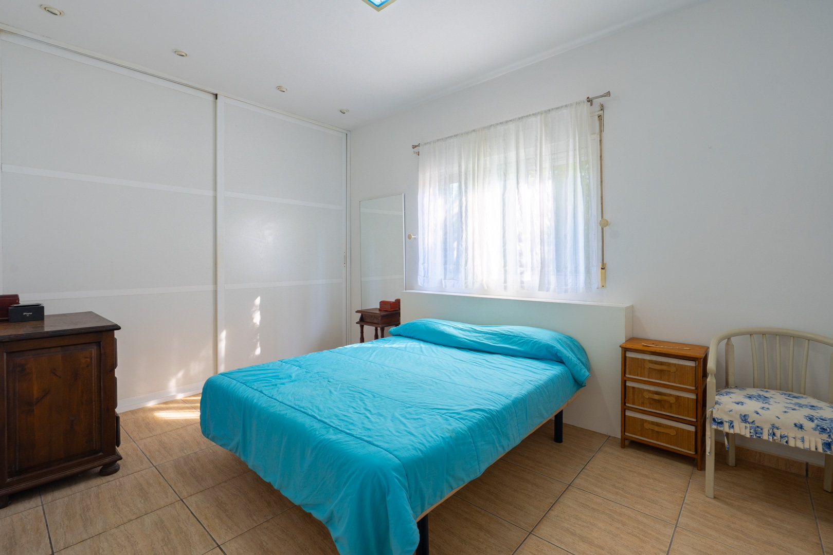 Dormitorio luminoso con armarios empotrados blancos, suelo de gres, ventana con cortinas ligeras y mobiliario auxiliar de madera clásica.