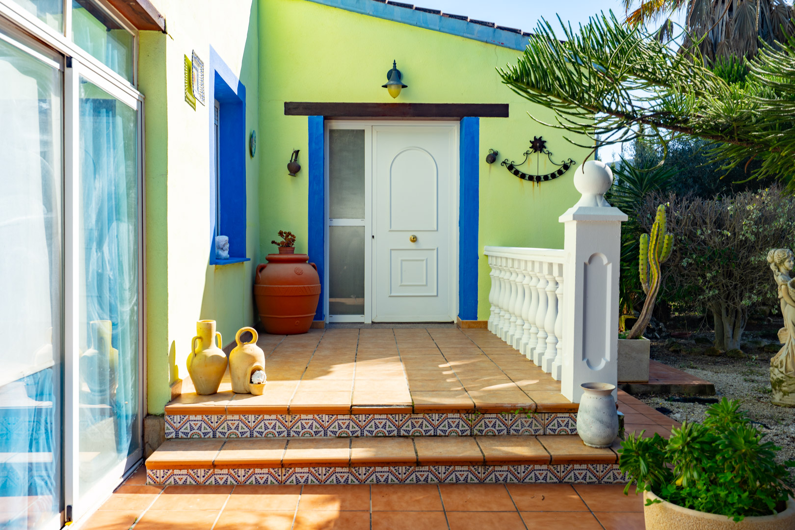 Entrada principal con puerta blanca, marcos azules y escalones con azulejos decorativos. La terraza de terracota incluye una balustrada blanca y elementos decorativos tradicionales.