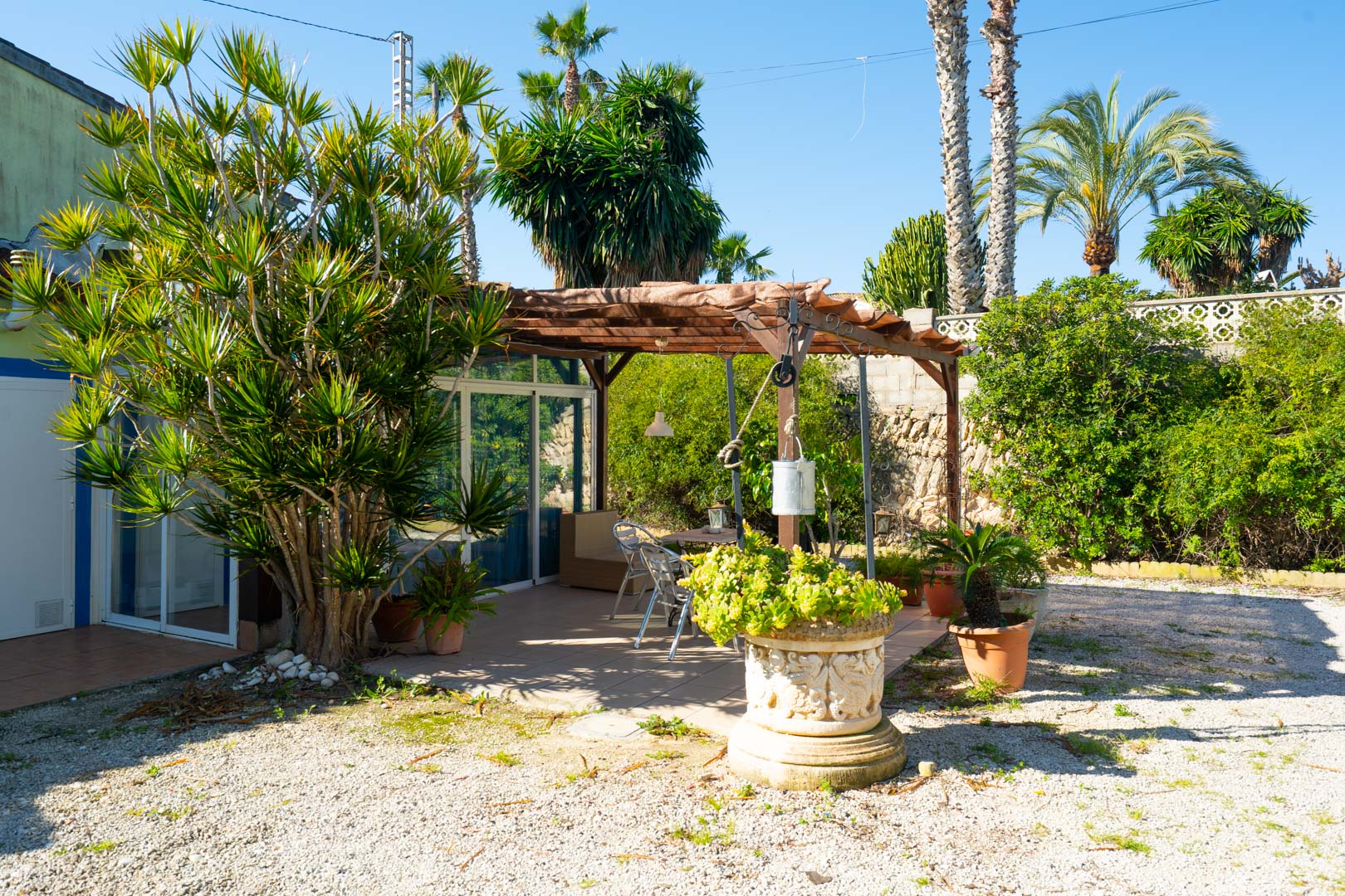Terraza exterior con pérgola de madera, zona de comedor y abundante vegetación mediterránea sobre suelo de grava y baldosas cerámicas.