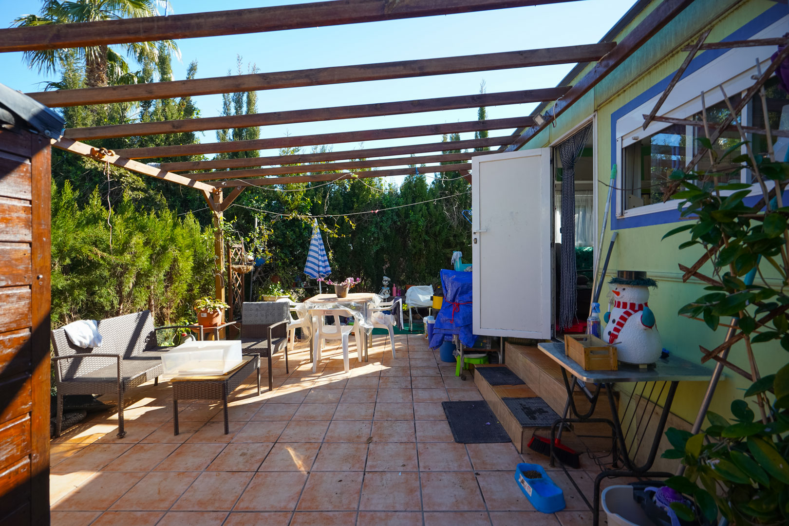 Terraza exterior de la vivienda prefabricada con suelo cerámico, pérgola de vigas de madera y zona de estar rodeada de vegetación privada.