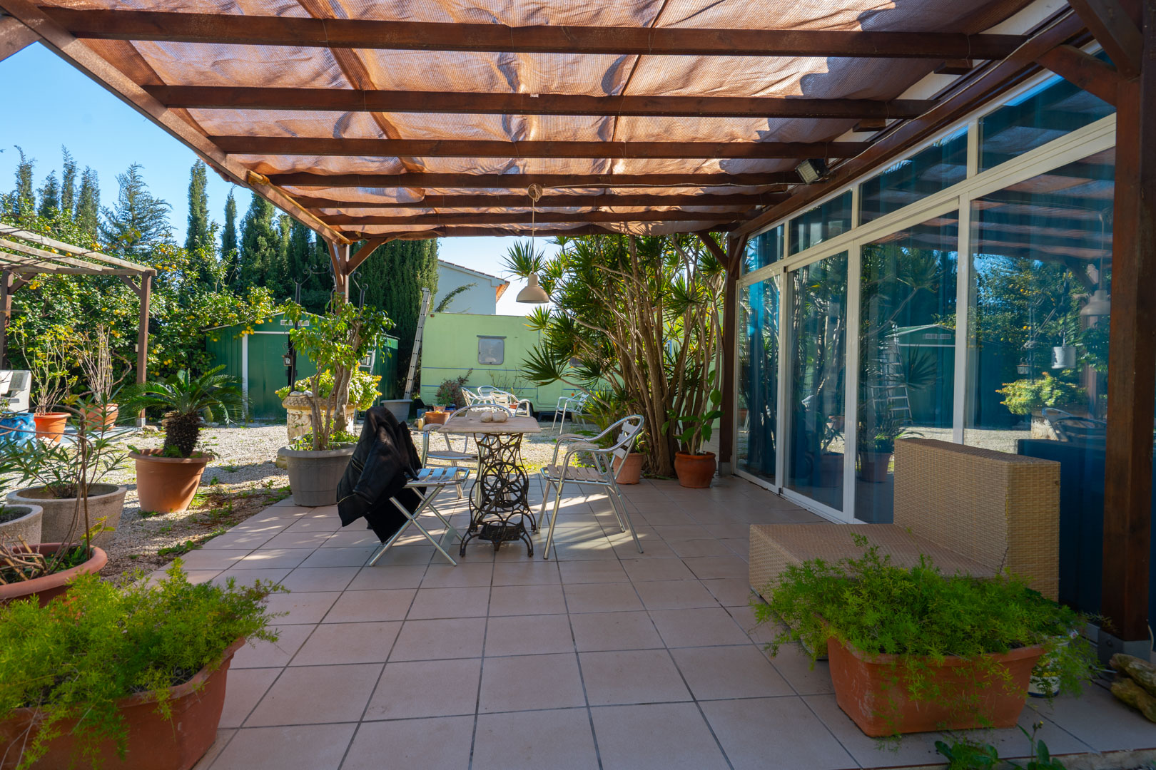 Terraza pavimentada con pérgola de madera y toldo de sombra, equipada con mobiliario de exterior y rodeada de plantas en macetas junto al jardín.