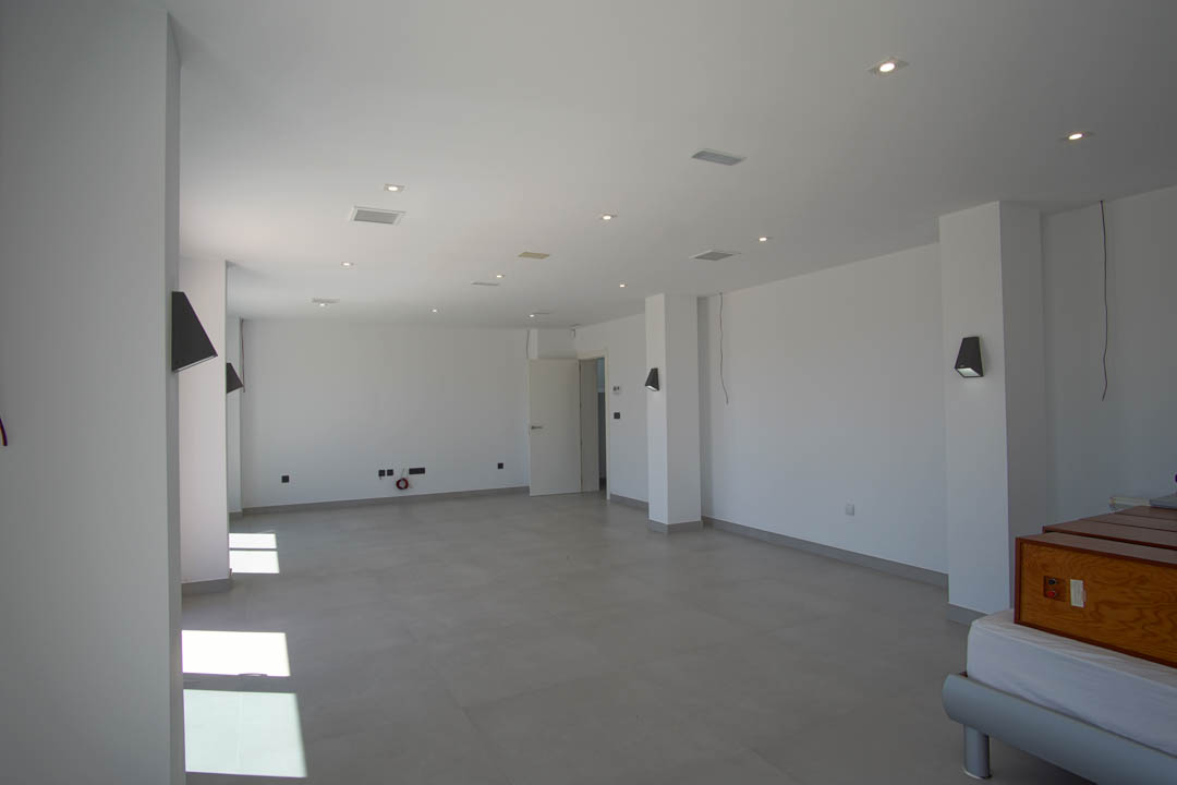 Amplia estancia diáfana con suelos de gres gris, paredes blancas, iluminación empotrada en el techo y apliques de pared modernos.