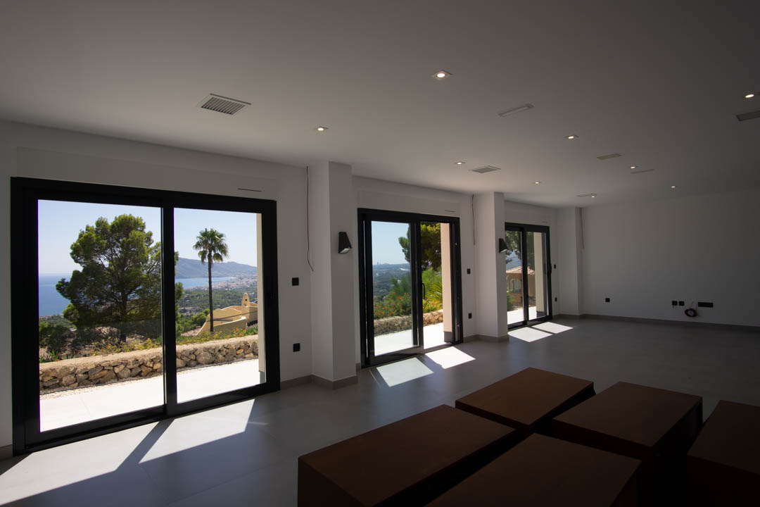 Amplio salón diáfano con ventanales de marco negro, suelos de gres gris y vistas panorámicas a la bahía de Altea desde la Sierra.