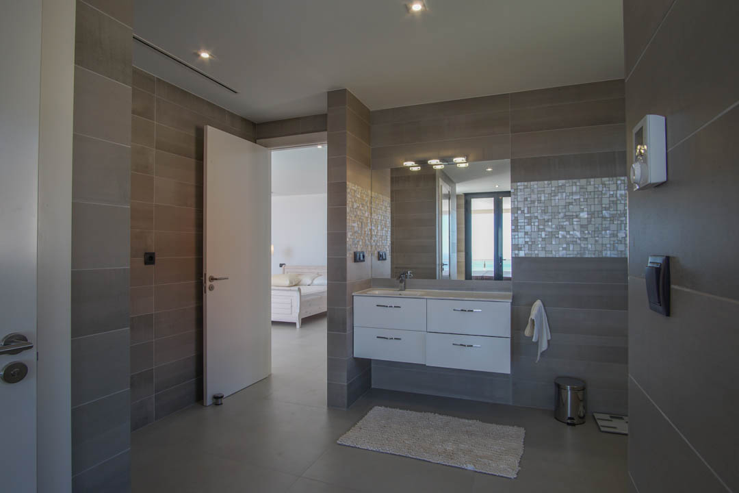 Baño de diseño con gres gris, lavabo suspendido blanco y detalles en mosaico. La estancia conecta visualmente con el dormitorio principal y el horizonte marino.