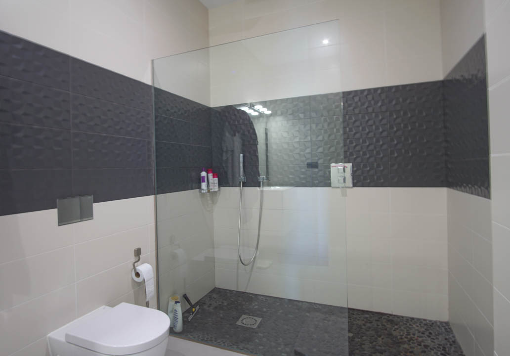 Baño moderno con ducha italiana de suelo de piedra, mampara de cristal fija y paredes revestidas con azulejos texturizados en gris oscuro y blanco.