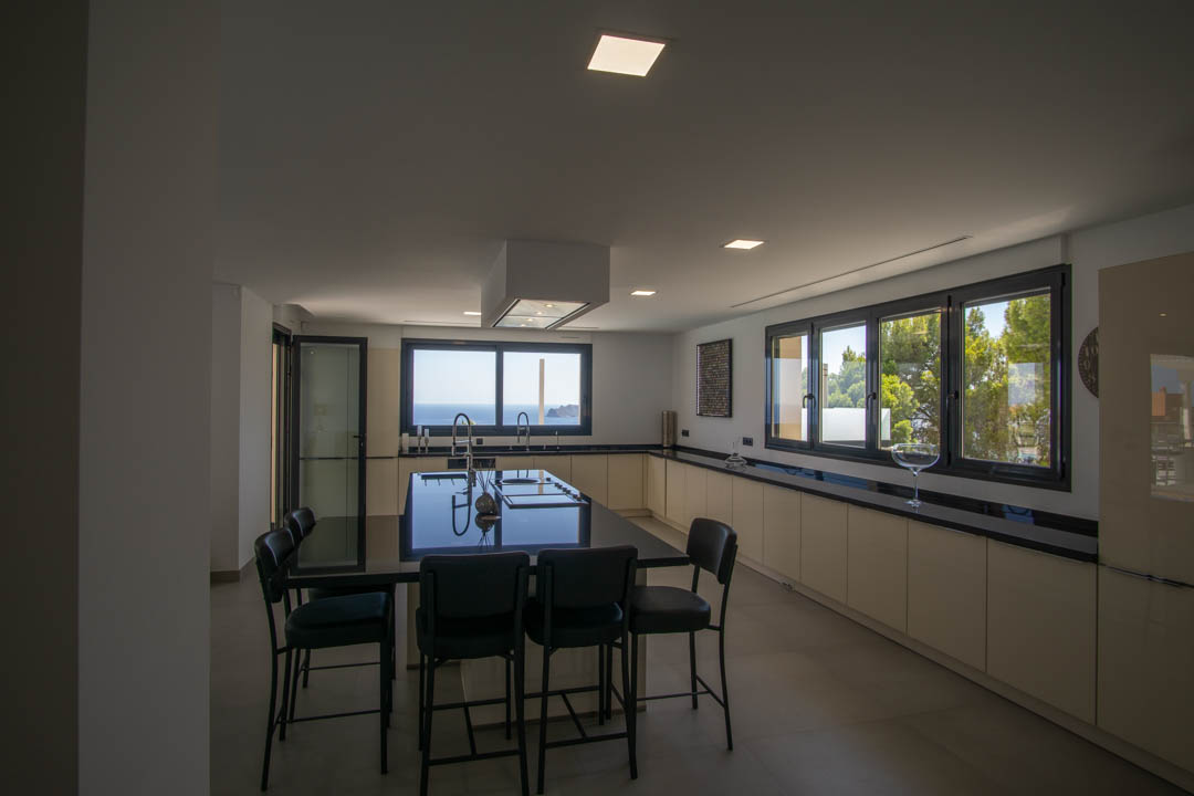 Cocina moderna con isla central de encimera negra, campana extractora suspendida y amplios ventanales con vistas al mar y al entorno natural de la Sierra de Altea.