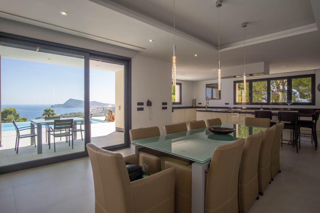 Comedor de concepto abierto con mesa de cristal y cocina integrada, conectado mediante amplios ventanales a la terraza con vistas panorámicas a la bahía.