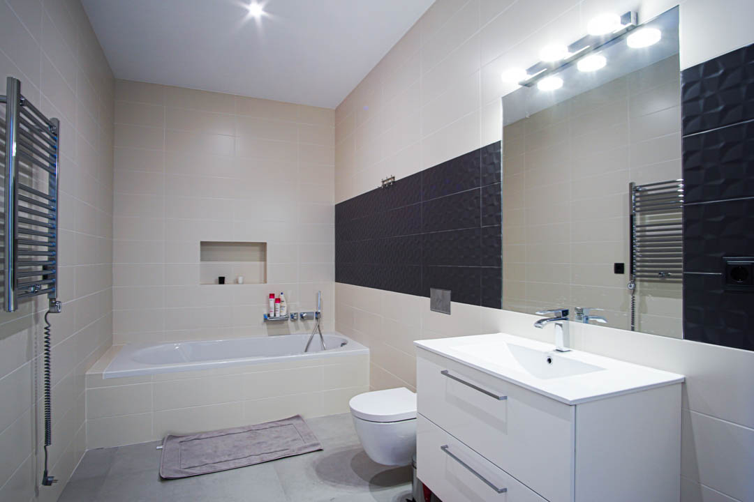 Cuarto de baño moderno con bañera encastrada, mueble de lavabo blanco, inodoro suspendido y paredes alicatadas en tonos beige con una franja de contraste oscura.