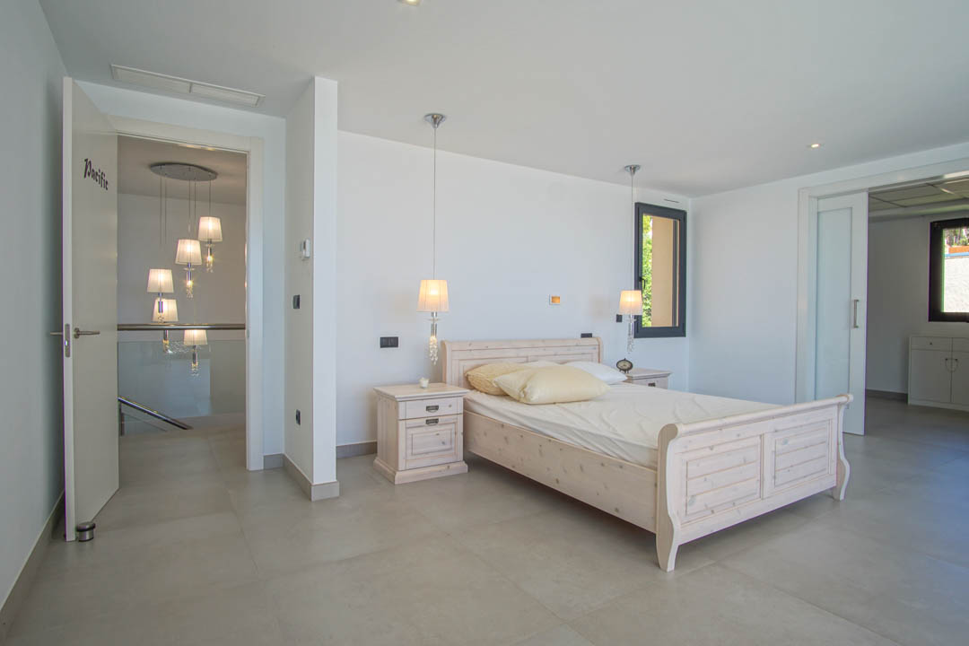 Dormitorio con cama de madera clara, mesitas a juego y lámparas colgantes. Suelo de gres porcelánico y acceso al pasillo con barandilla de cristal y lámparas de diseño.
