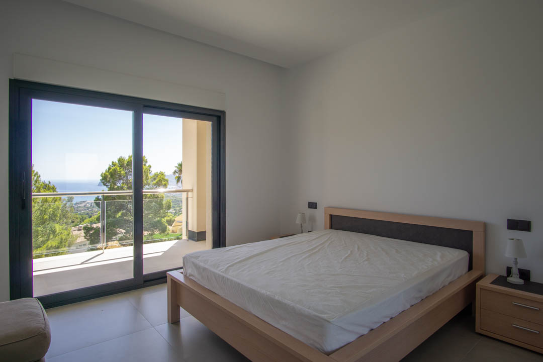 Dormitorio luminoso con estructura de cama en madera clara, acceso directo a un balcón privado y vistas despejadas hacia la costa y la vegetación mediterránea.