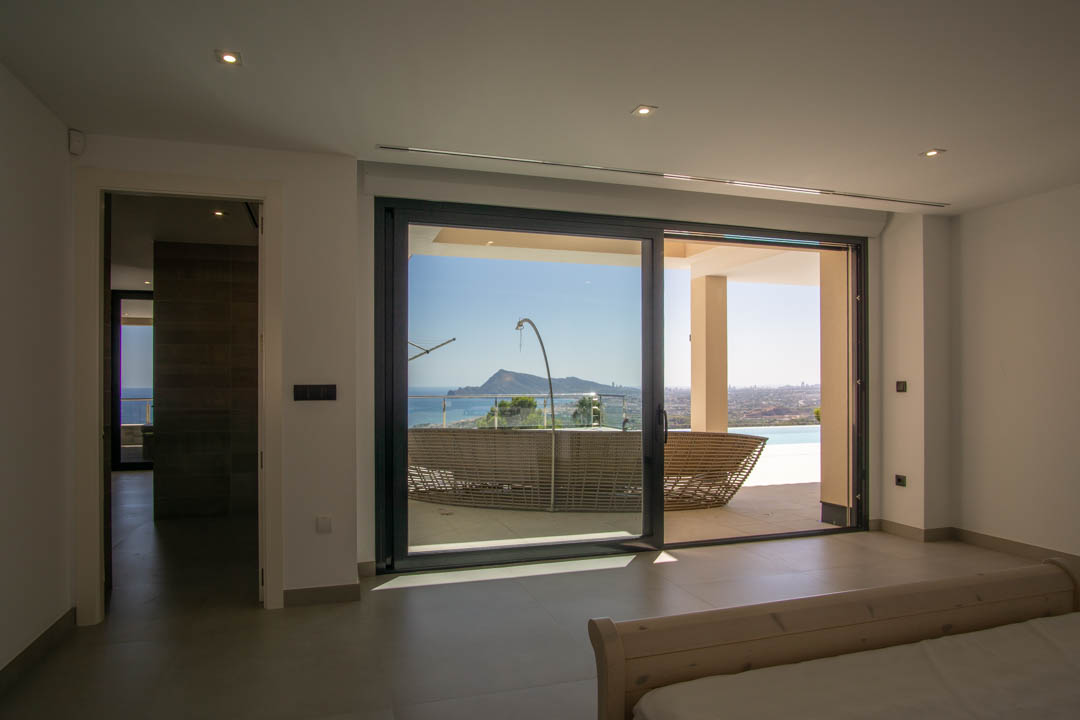 Dormitorio principal con acceso directo a la terraza mediante grandes ventanales, ofreciendo vistas panorámicas al mar y a la montaña bajo iluminación empotrada.