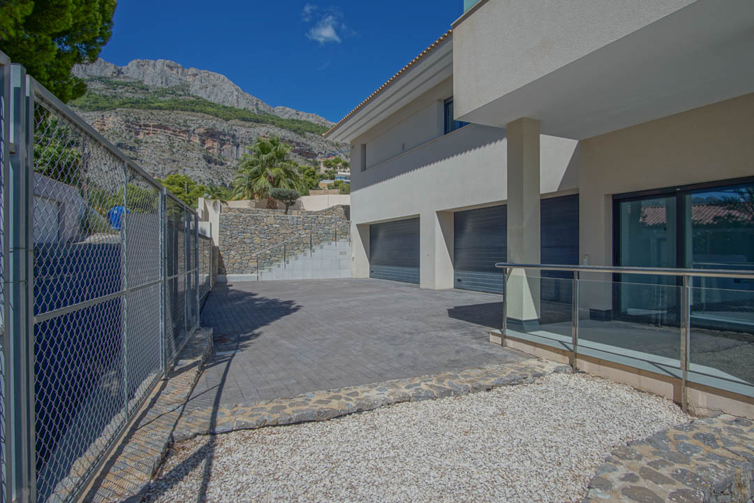 Entrada exterior con acceso a garaje triple, zona pavimentada, muros de piedra natural y vistas a la Sierra de Altea bajo un cielo despejado.