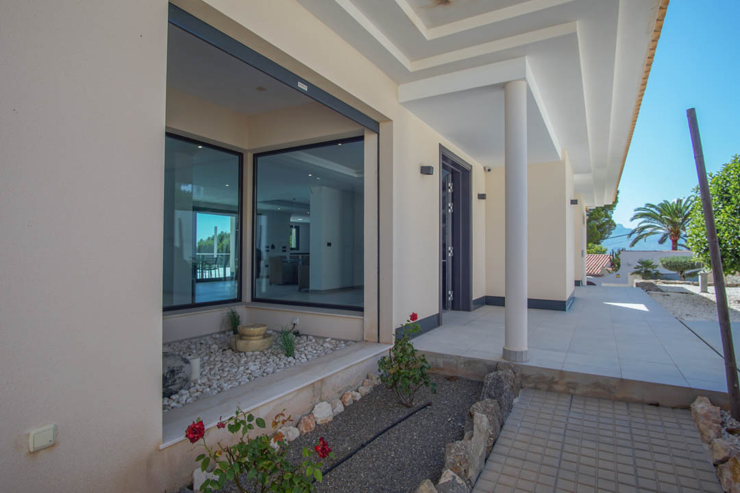 Entrada principal con porche cubierto, pilares blancos y grandes ventanales. Incluye un jardín decorativo con piedras blancas y vistas al entorno natural de Altea.