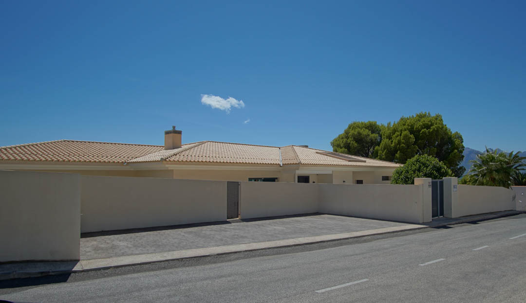 Fachada exterior de la villa con muro perimetral de seguridad, zona de aparcamiento pavimentada y acceso privado bajo un cielo despejado.