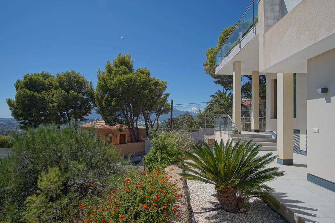 Fachada exterior moderna con columnas blancas, barandillas de cristal y zona ajardinada con grava, palmeras y vegetación mediterránea bajo un cielo despejado.