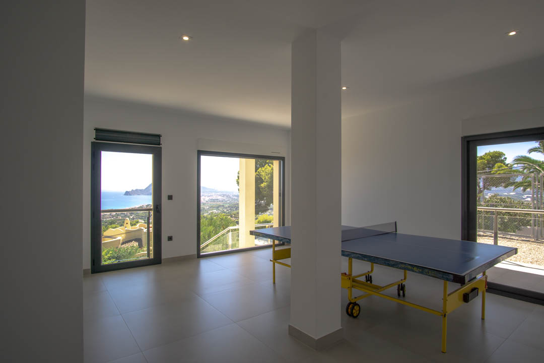 Sala de juegos con mesa de ping-pong, suelos de gres gris y amplios ventanales que ofrecen vistas panorámicas al mar Mediterráneo y la costa de Altea.