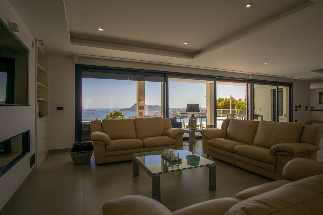 Salón espacioso con sofás de cuero, iluminación empotrada y grandes ventanales que ofrecen vistas panorámicas al mar y a la bahía de Altea.