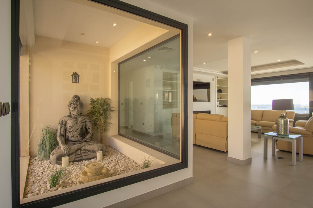 Salón moderno con jardín zen interior acristalado que incluye una estatua de Buda sobre cantos rodados blancos, junto a una zona de estar con sofás de cuero y vistas al mar.