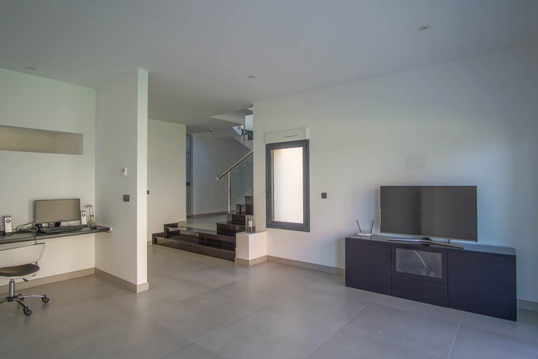 Salón secundario con zona de despacho integrada, suelos de gres gris y acceso a la planta superior mediante escalera de diseño con barandilla de cristal.