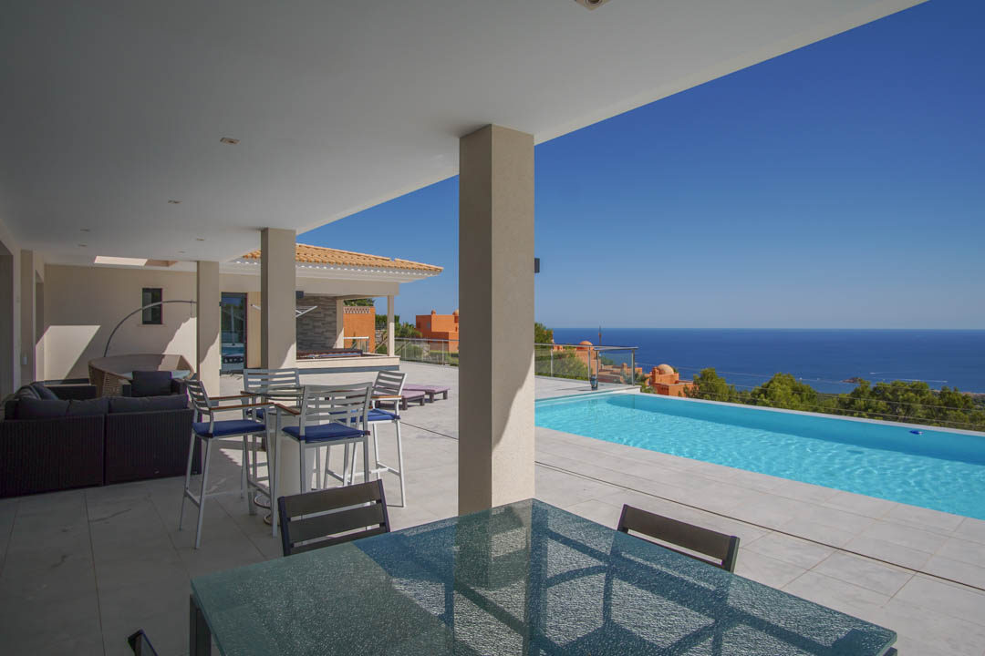 Terraza cubierta con suelos de piedra clara, zona de comedor exterior y piscina infinita con vistas panorámicas al mar Mediterráneo en Altea.