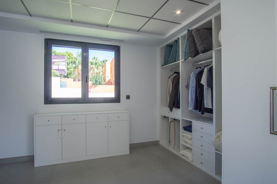 Vestidor con mobiliario a medida en blanco, cajoneras integradas y suelo cerámico gris. Dispone de ventana con marco oscuro que aporta luz natural y vistas al exterior.