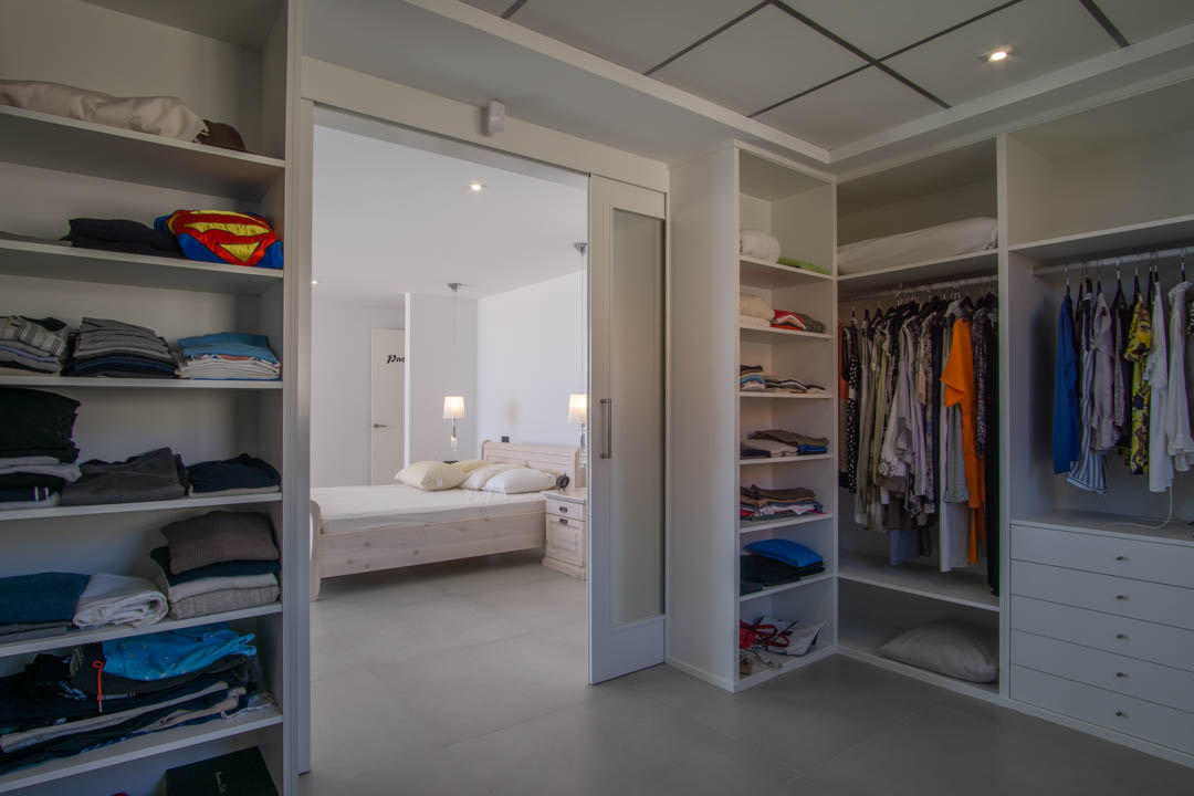 Vestidor de 12m2 con estanterías modulares blancas y cajonera, conectado al dormitorio principal mediante una puerta corredera de cristal translúcido.