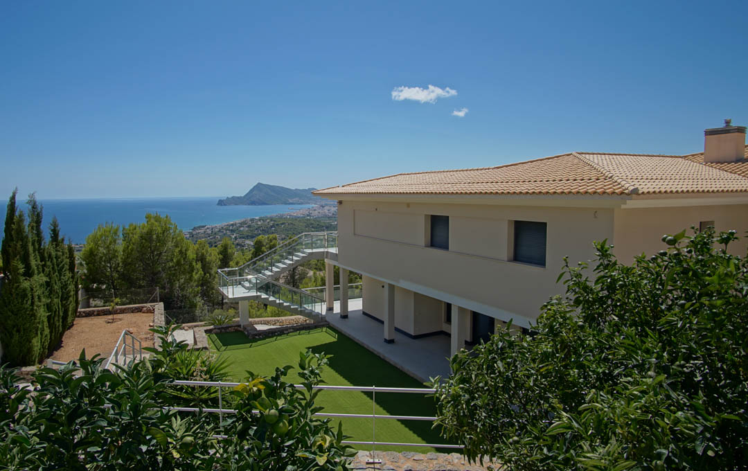 Vista exterior de la villa con jardín de césped artificial, árboles frutales y escalera de cristal, ofreciendo vistas panorámicas a la bahía de Altea y el Mediterráneo.