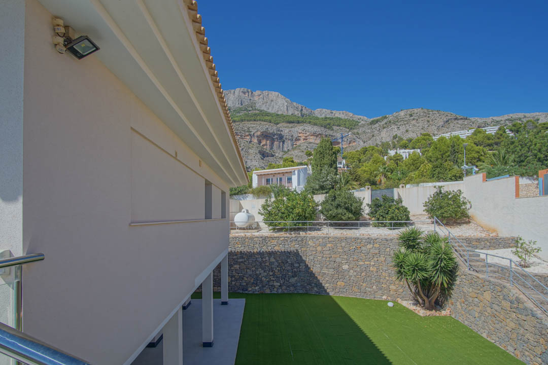Vista exterior del jardín con césped artificial, muros de piedra natural y zona de árboles frutales, enmarcada por las montañas de la Sierra de Altea.