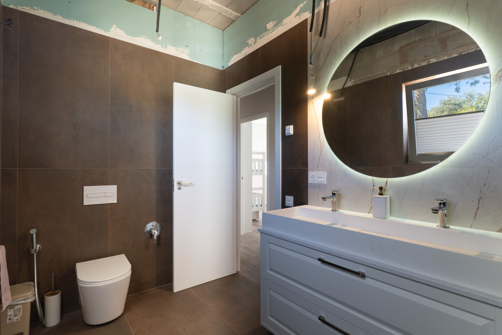 Baño contemporáneo con lavabo doble, espejo circular retroiluminado y azulejos en tonos tierra. Se aprecia el inodoro suspendido y los acabados técnicos en proceso de finalización.