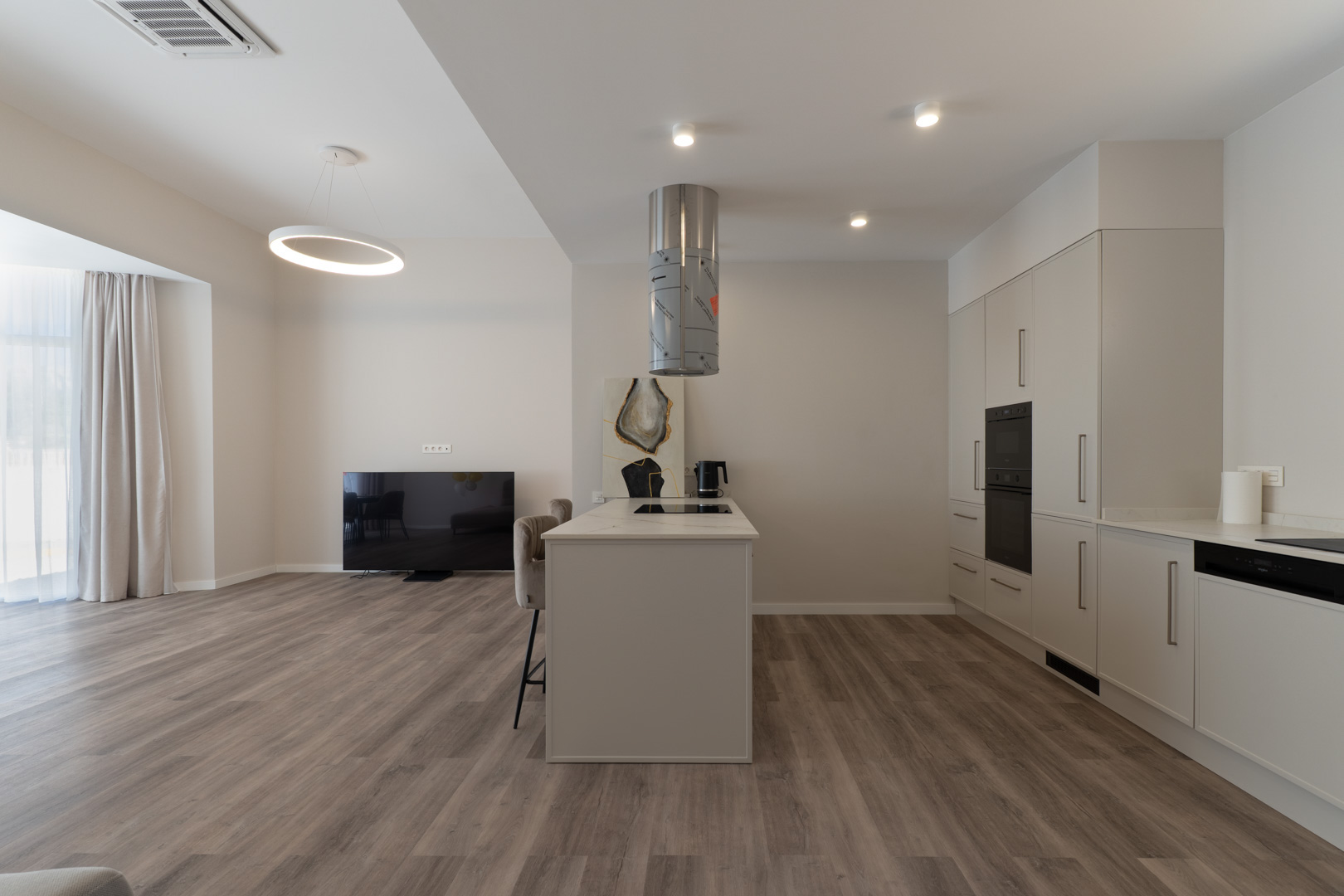 Cocina de concepto abierto con isla central, encimera de efecto mármol y campana cilíndrica, integrada en un salón con suelos de madera laminada y tonos neutros.