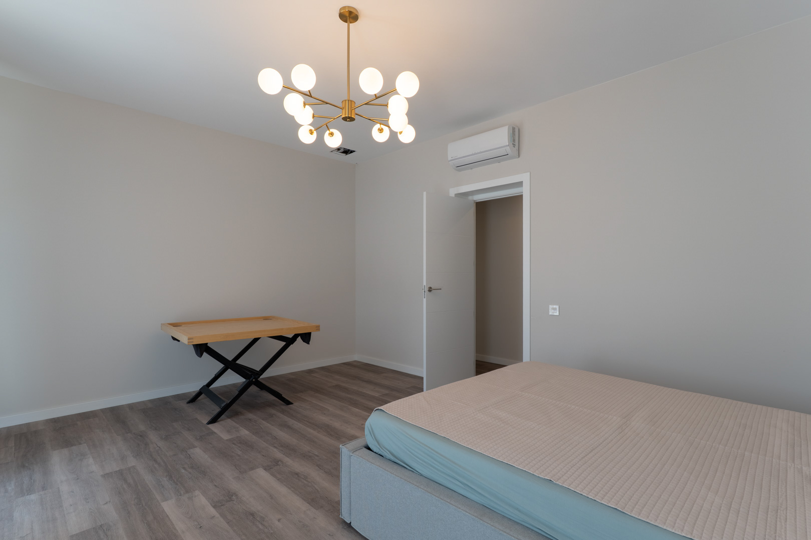 Dormitorio contemporáneo con suelos de tarima gris, paredes neutras y una lámpara de diseño en dorado. Incluye aire acondicionado y acceso directo a través de una puerta blanca.