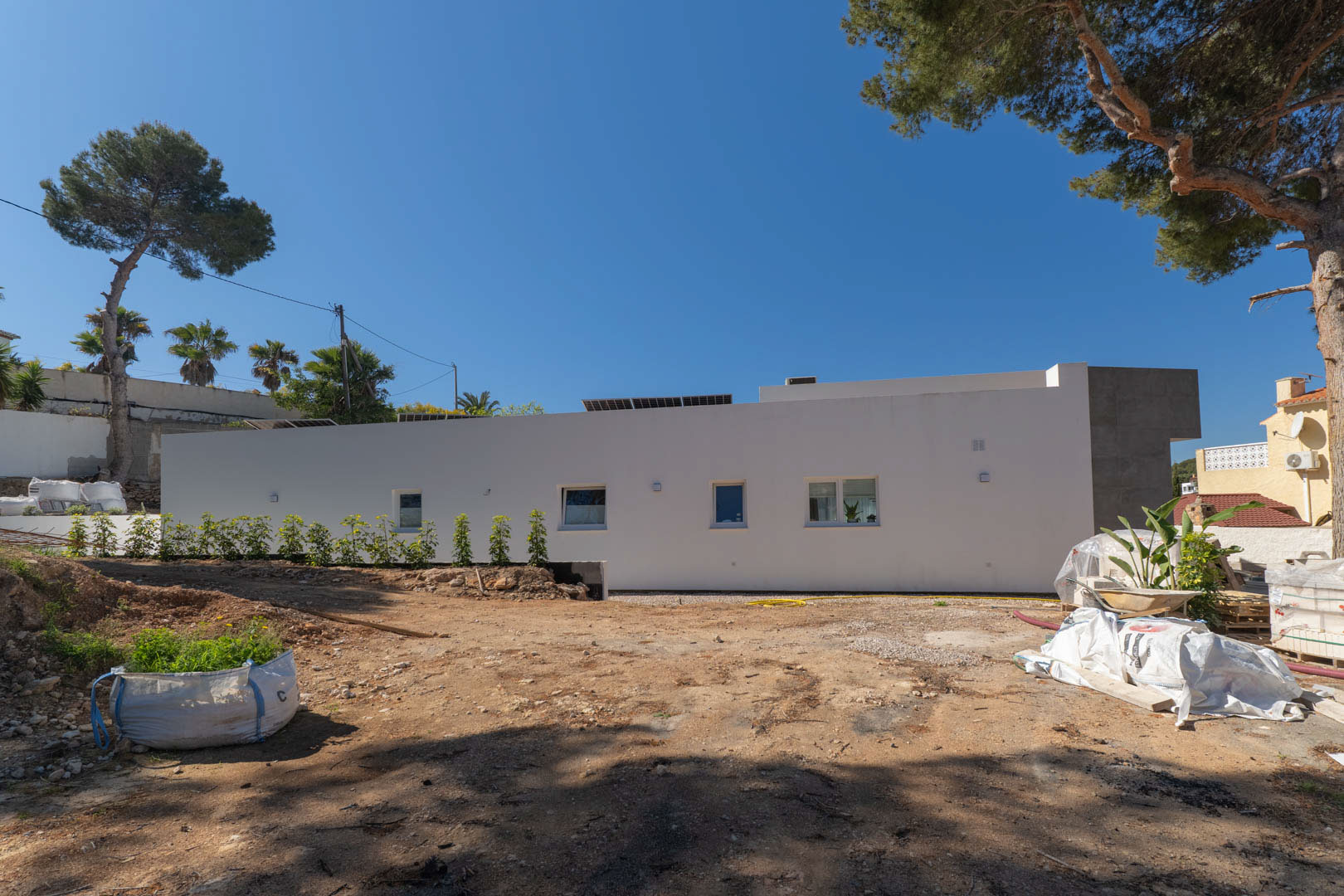 Fachada lateral de la villa minimalista con paneles solares, mostrando el terreno exterior en fase de preparación y la integración de pinos maduros bajo un cielo despejado.