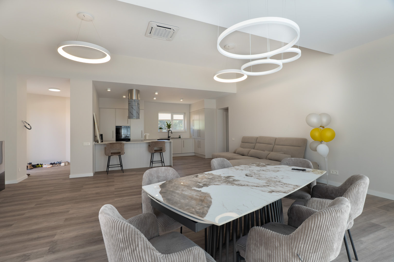 Salón-comedor diáfano con mesa de mármol, sillas tapizadas y cocina minimalista integrada con isla. Destacan las luminarias LED circulares y el suelo de acabado madera.