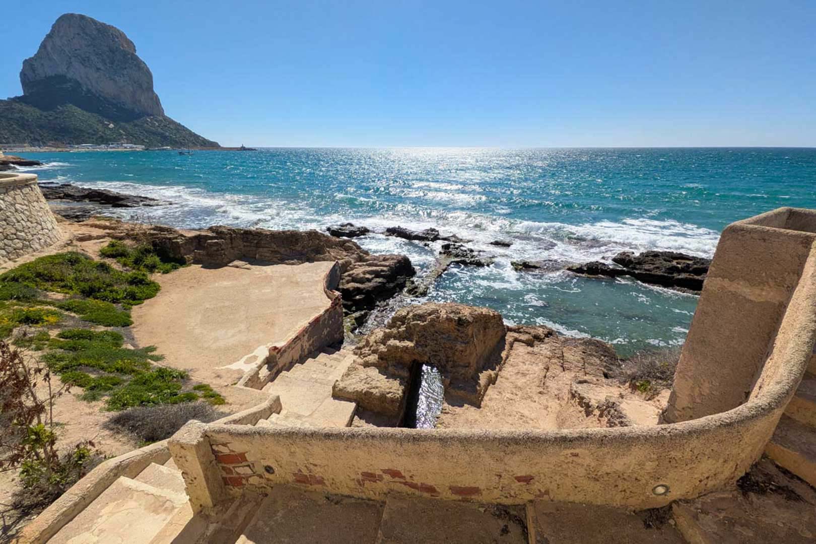 Vistas panorámicas al Peñón de Ifach desde una terraza escalonada de piedra, con acceso directo a una cala de aguas turquesas y entorno natural.