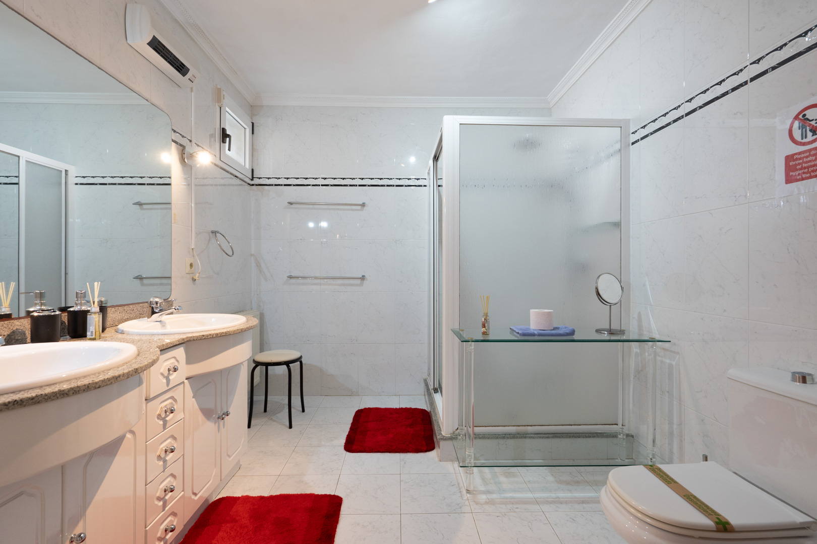 Baño completo con lavabo doble sobre encimera de granito, espejo de gran formato, ducha con mampara translúcida y climatización individual mediante split.